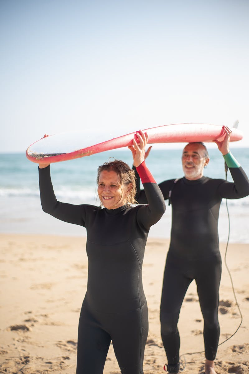 Elderly couple in wetsuits having fun with a surfboard on a sunny beach in Portugal